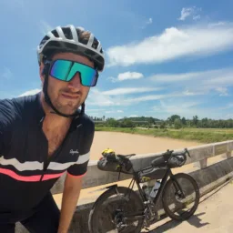 Close up of Matt wearing cycle helmet and sunglasses standing on dirt track with blue sky