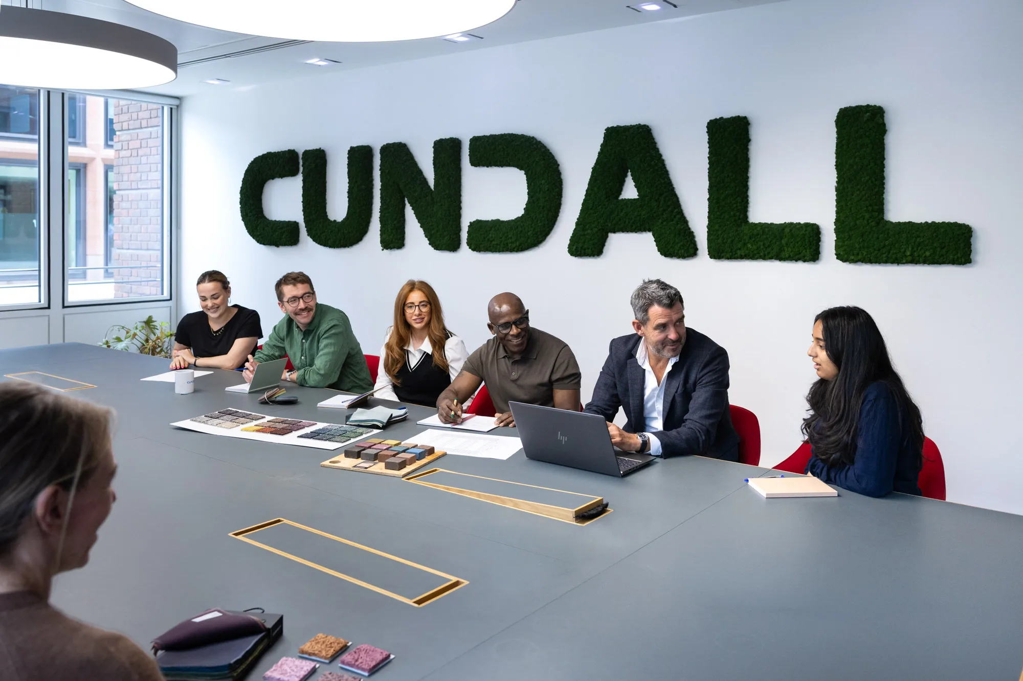 People seated around a large meeting table beneath a moss-covered Cundall wall sign, with laptops, notebooks and material samples displayed in a bright office interior.