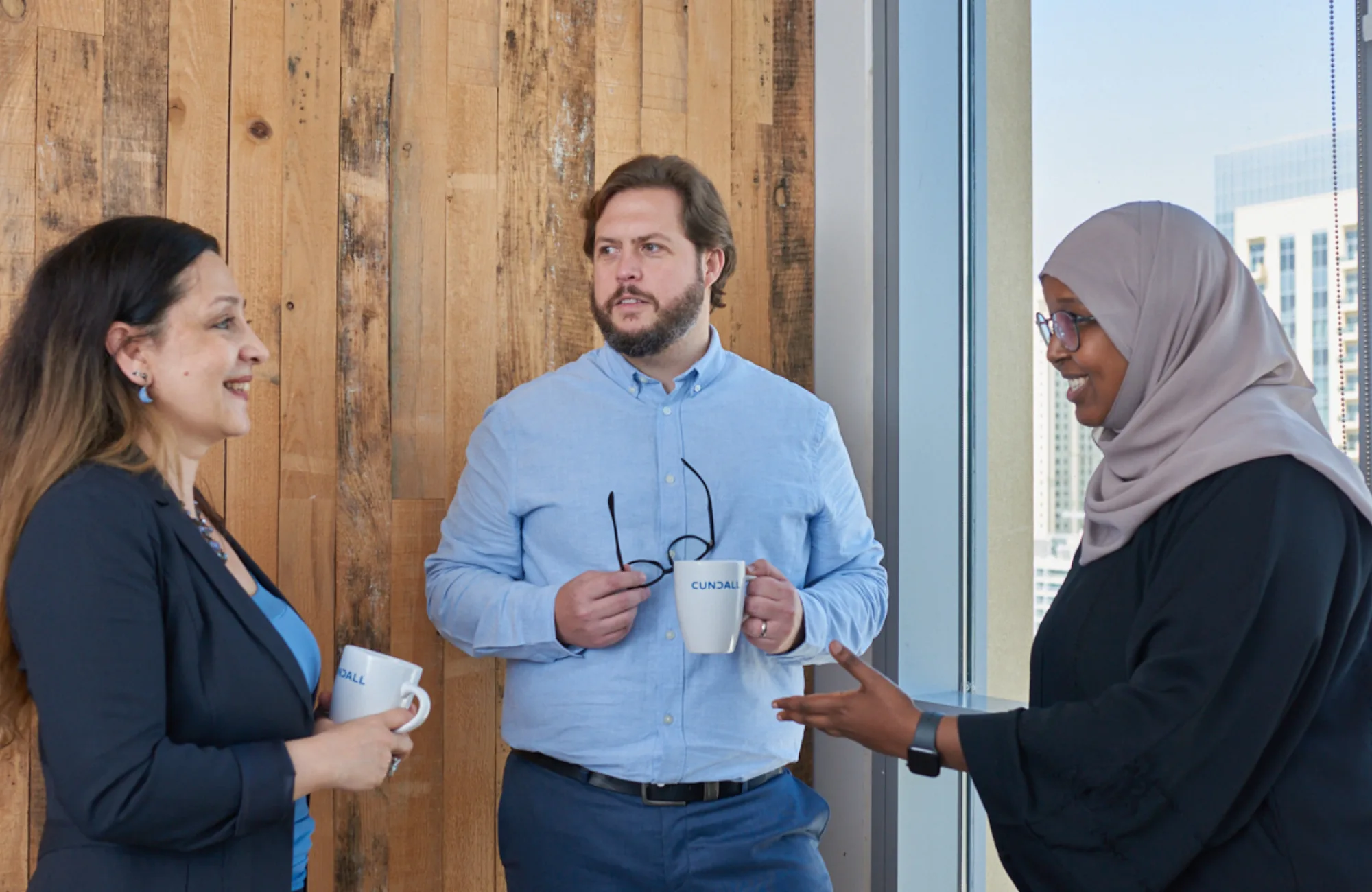 3 people standing having a conversation against a wood-panelled wall and a light window