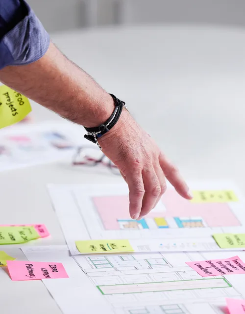 A person’s hand reaches over a table covered with architectural drawings and scattered sticky notes in various colors, while another hand rests nearby at the edge of the table.