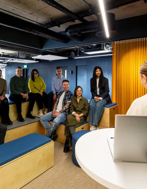 A presenter behind a podium in front of a large screen and a small audience on wooden box theatre style seating in the office space.