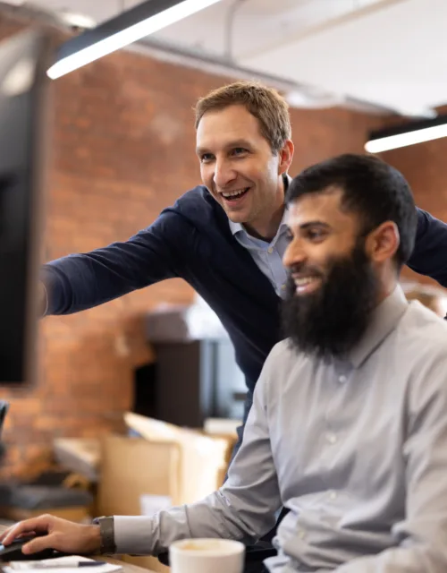 Staff in office one at a desk and the other standing behind looking at screen
