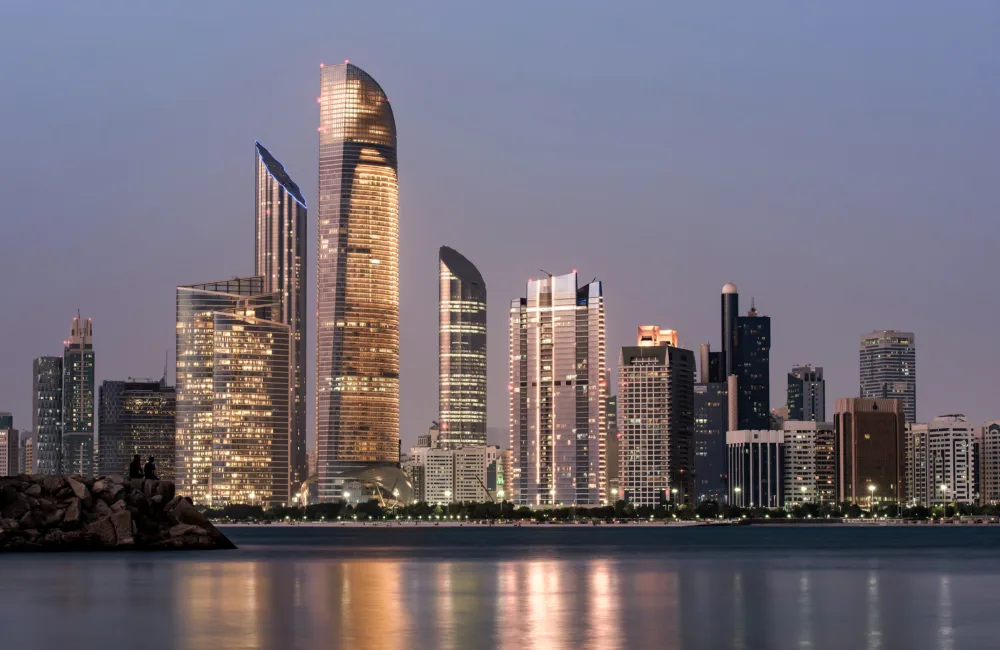 A waterfront city skyline at dusk featuring illuminated high-rise buildings with varied shapes, reflected in calm water, with a rocky shoreline in the foreground and a clear evening sky above.