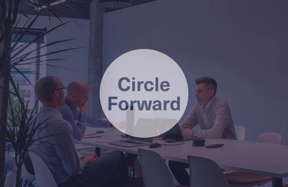 Three people seated around a table with laptops in a commercial office interior featuring exposed concrete columns, ductwork and indoor planting.