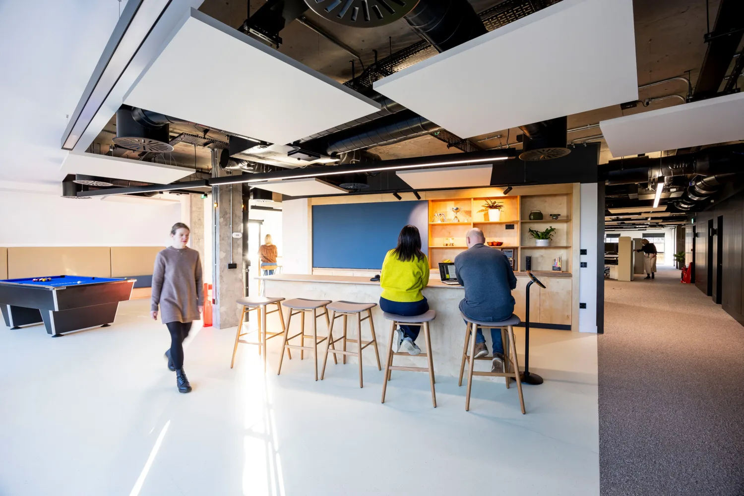 Open-plan office interior with a central counter, seating, pool table, exposed ceiling services and people using the workspace