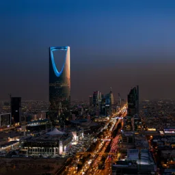 Night cityscape with illuminated skyscrapers and dense low-rise buildings; a tall tower with a curved opening glows blue at center. A wide highway in the foreground shows heavy traffic with red and white lights stretching into the distance.