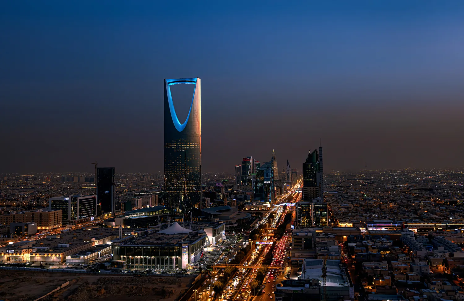 Night cityscape with illuminated skyscrapers and dense low-rise buildings; a tall tower with a curved opening glows blue at center. A wide highway in the foreground shows heavy traffic with red and white lights stretching into the distance.