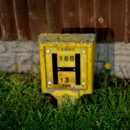Yellow rectangular hydrant marker set in grass, weathered with chipped paint and lichen on top. Front panel shows a large black “H,” numbers “180” and “13,” and smaller number “18502” above. Corrugated metal fence and low concrete edging in background.