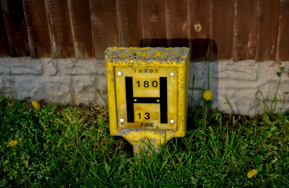 Yellow rectangular hydrant marker set in grass, weathered with chipped paint and lichen on top. Front panel shows a large black “H,” numbers “180” and “13,” and smaller number “18502” above. Corrugated metal fence and low concrete edging in background.