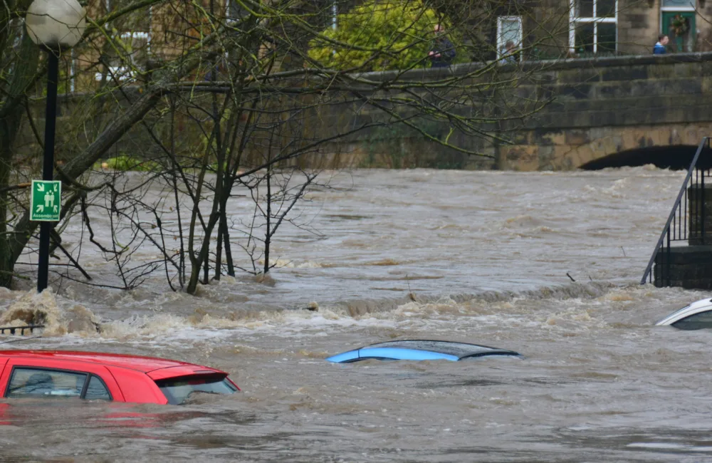 A flooded river with a concrete bridge in the background and the rooves of 3 cars visible above the water level.