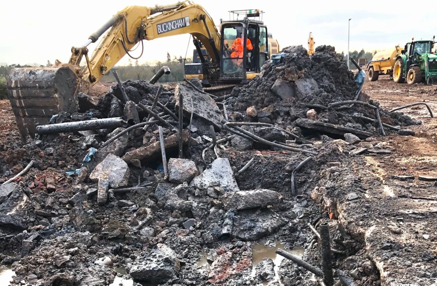A yellow excavator labeled "BUCKINGHAM" digs through rubble and debris, including broken concrete and metal rods. Two workers in orange safety gear are inside the cab. Construction vehicles are visible in the background.