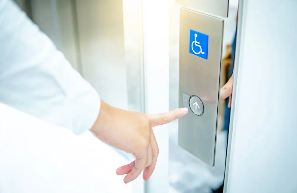Close-up of a hand pressing an elevator call button on a stainless steel panel. A blue wheelchair accessibility symbol is displayed above the round button, next to an open lift door.