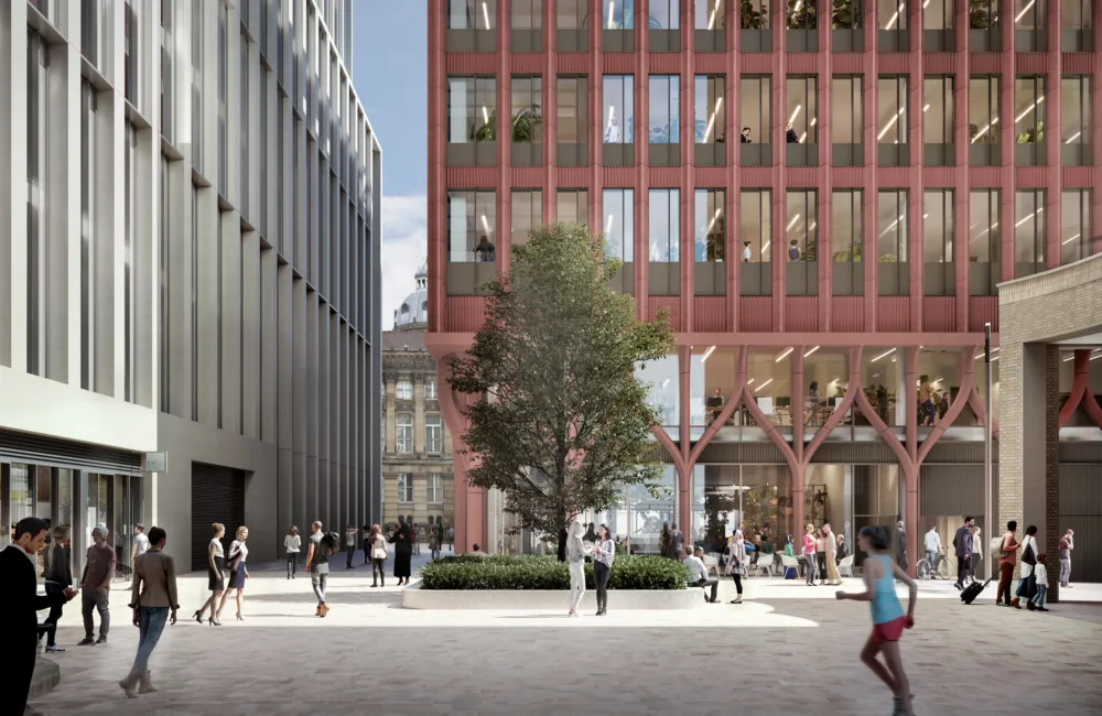 Modern urban plaza with a large central tree surrounded by benches. People are walking and interacting. A red building with large windows is on the right; a grey building is on the left.