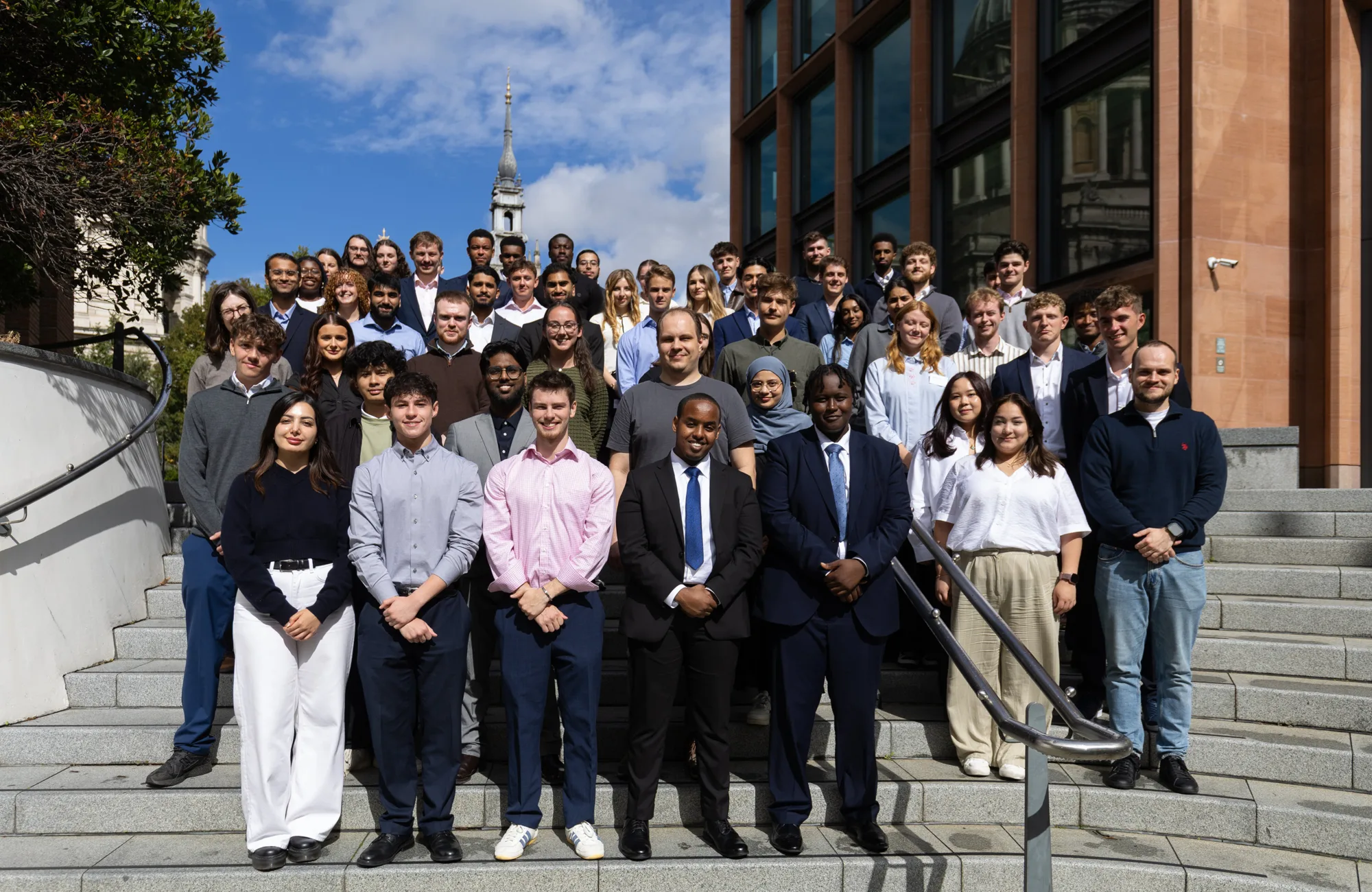 Large group of people standing on outdoor steps between office buildings in a city setting.