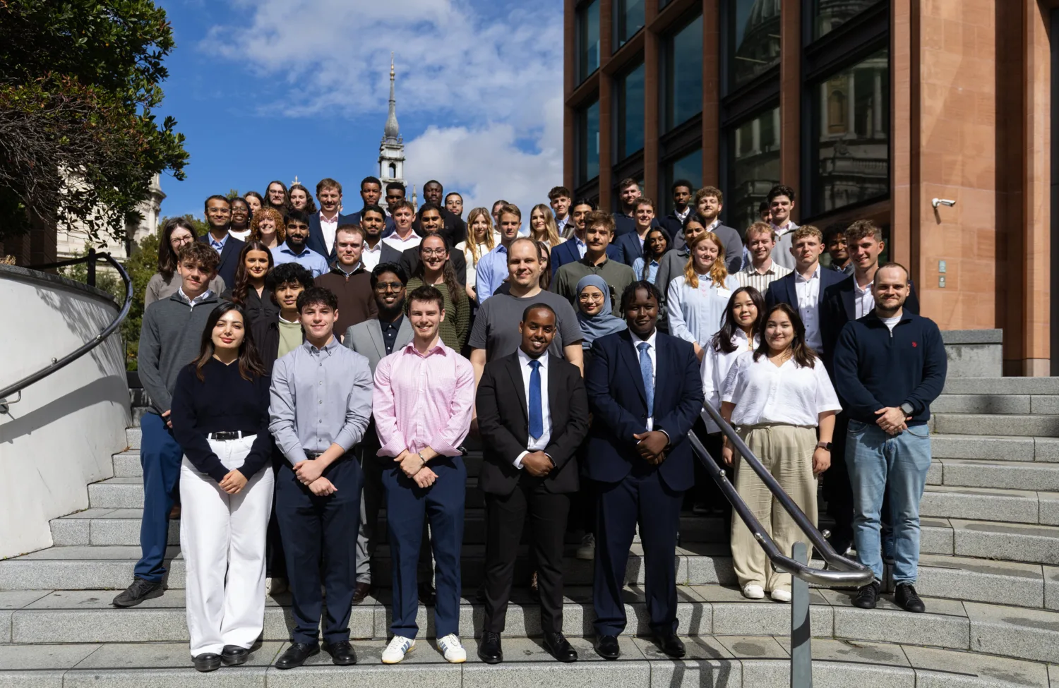 Large group of people standing on outdoor steps between office buildings in a city setting.
