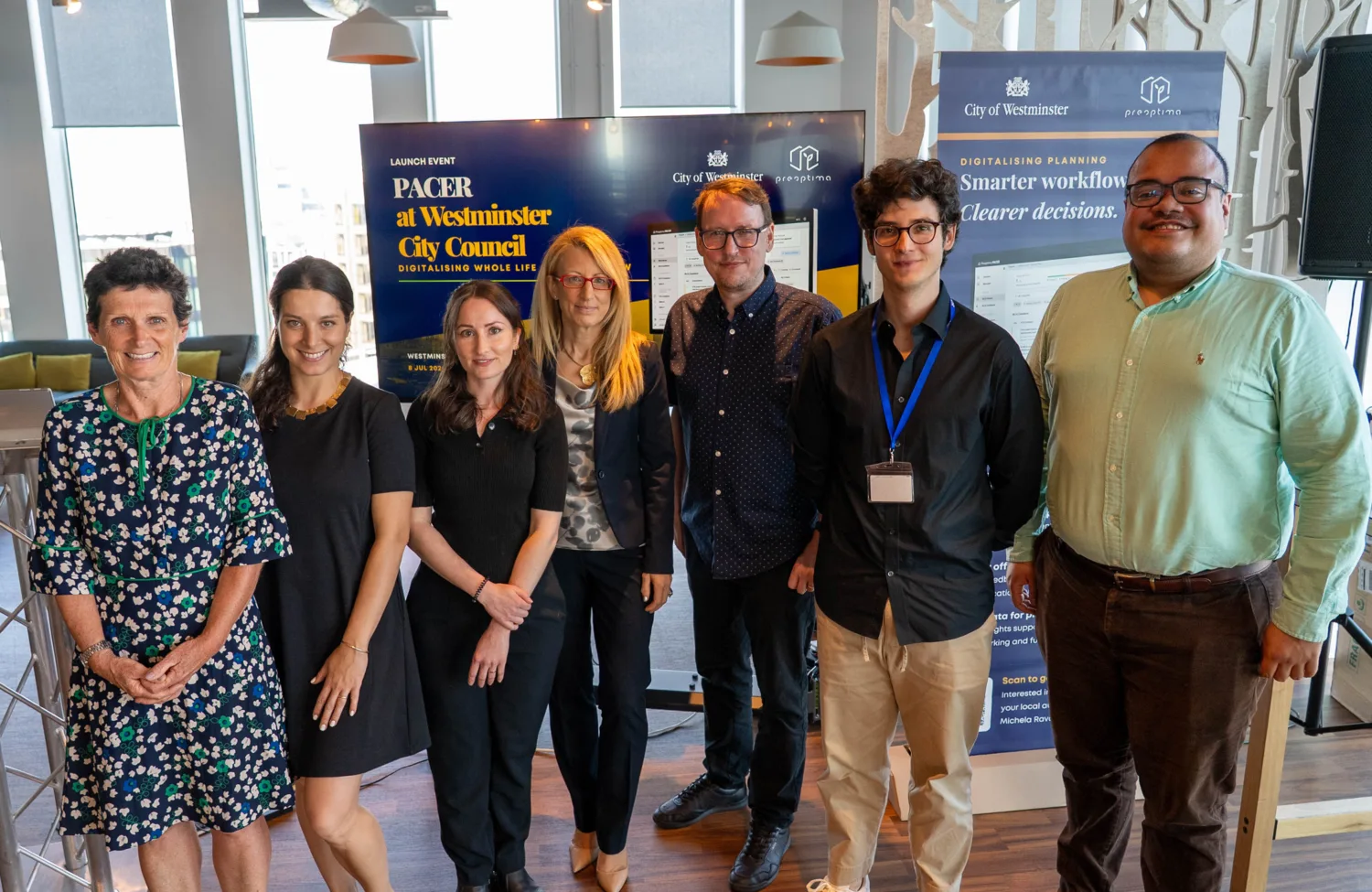 Group of people standing in front of PACER launch event display screens and branded banners in an interior space.