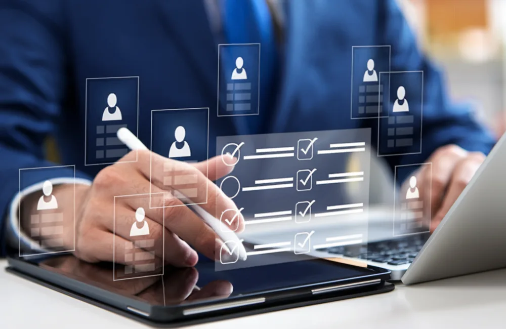 A person in a suit uses a stylus on a tablet beside an open laptop, with transparent digital icons showing user profiles and checklists floating above the devices.