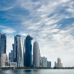 Modern waterfront skyline featuring multiple high-rise office and residential towers with glass façades along a shoreline. Distinctive buildings of varying shapes and heights stand beside calm water under a sky filled with layered white clouds. No people or visible text are present.