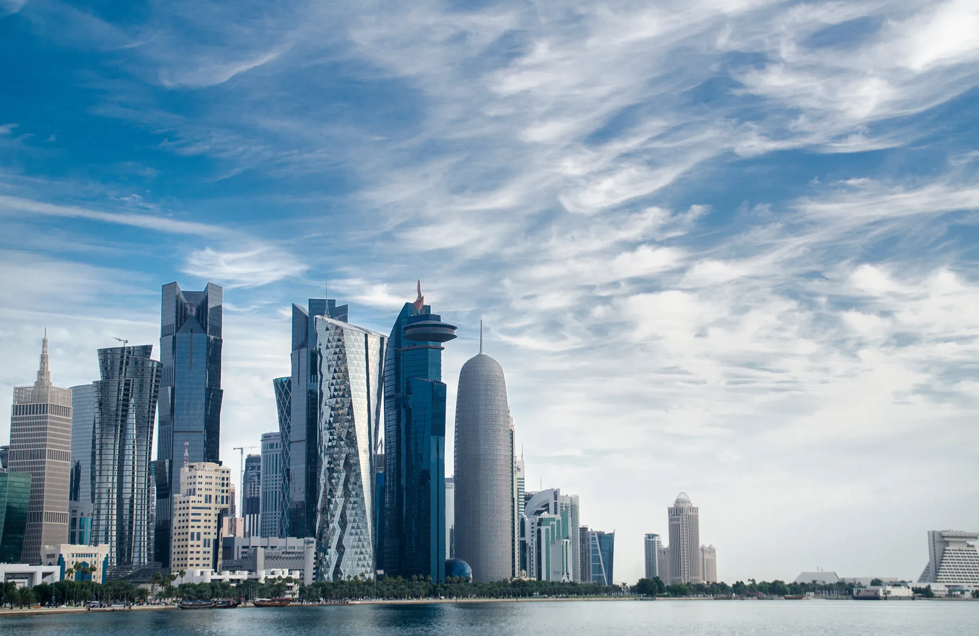 Modern waterfront skyline featuring multiple high-rise office and residential towers with glass façades along a shoreline. Distinctive buildings of varying shapes and heights stand beside calm water under a sky filled with layered white clouds. No people or visible text are present.