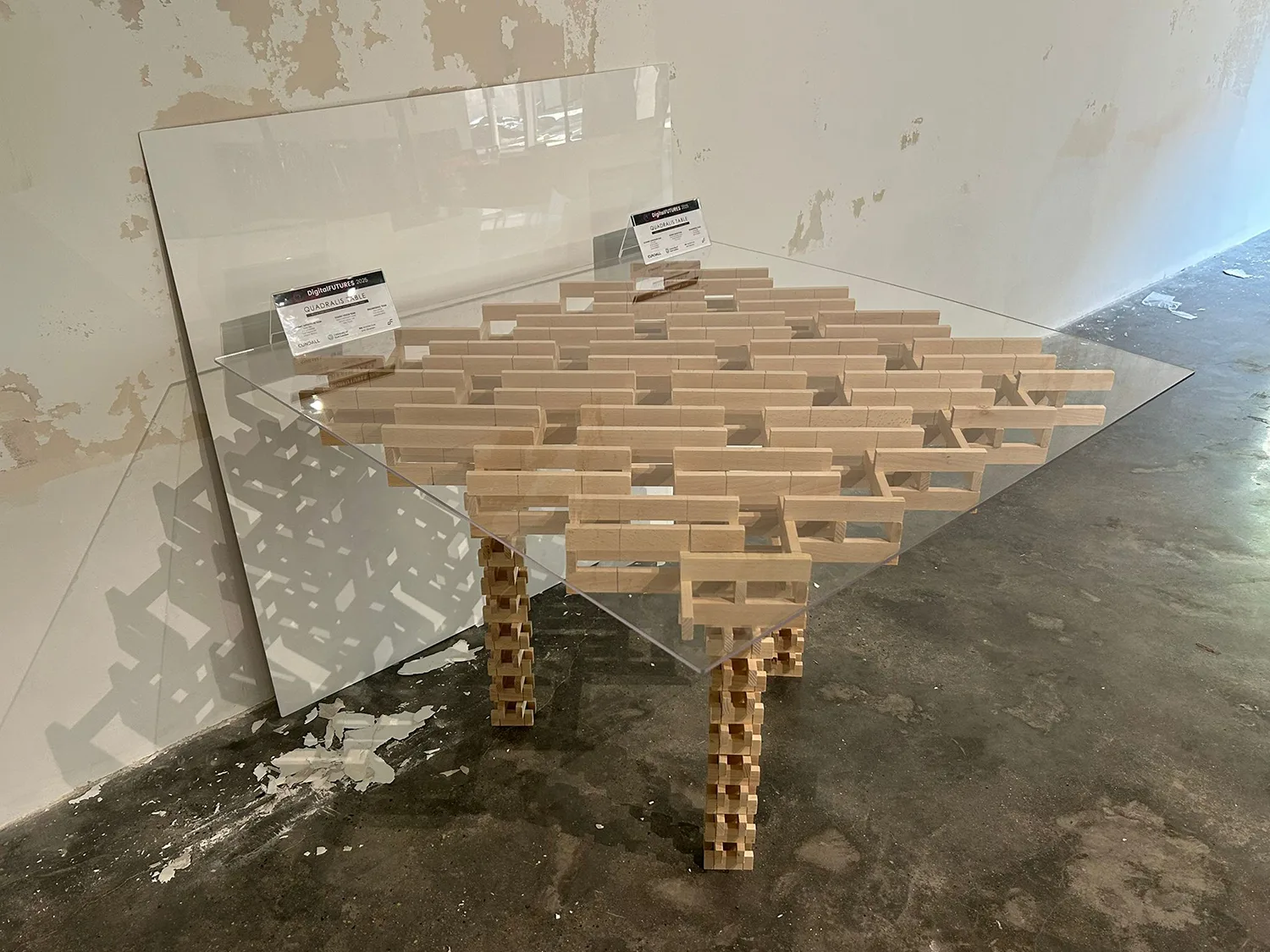 A glass table supported by a wooden block structure forming its base and legs; two small signs with partially legible text rest on top; background includes a peeled wall and concrete floor.