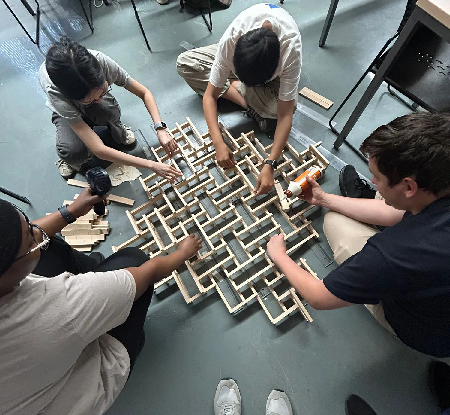 Four people seated on the floor assembling a lattice structure with small wooden sticks; one holds a glue bottle, another uses a drill, and two arrange sticks.
