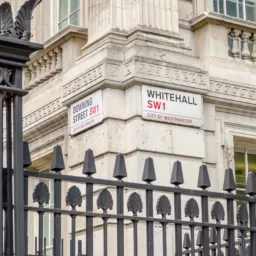 Street signs for Whitehall SW1 and Downing Street SW1 on a stone building corner in Westminster, with black iron railings in the foreground.