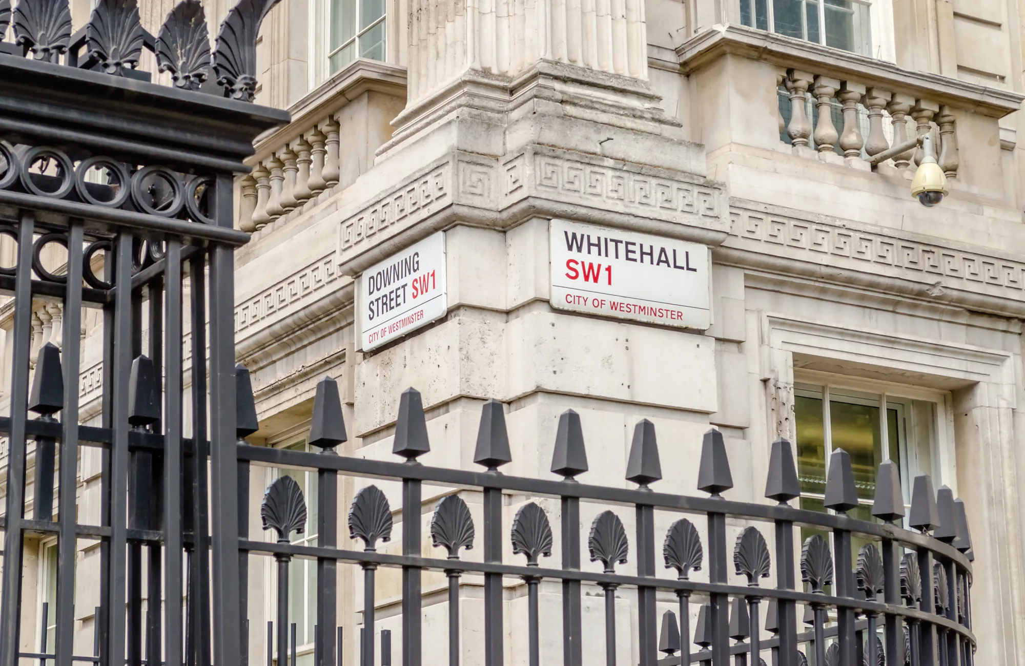 Street signs for Whitehall SW1 and Downing Street SW1 on a stone building corner in Westminster, with black iron railings in the foreground.