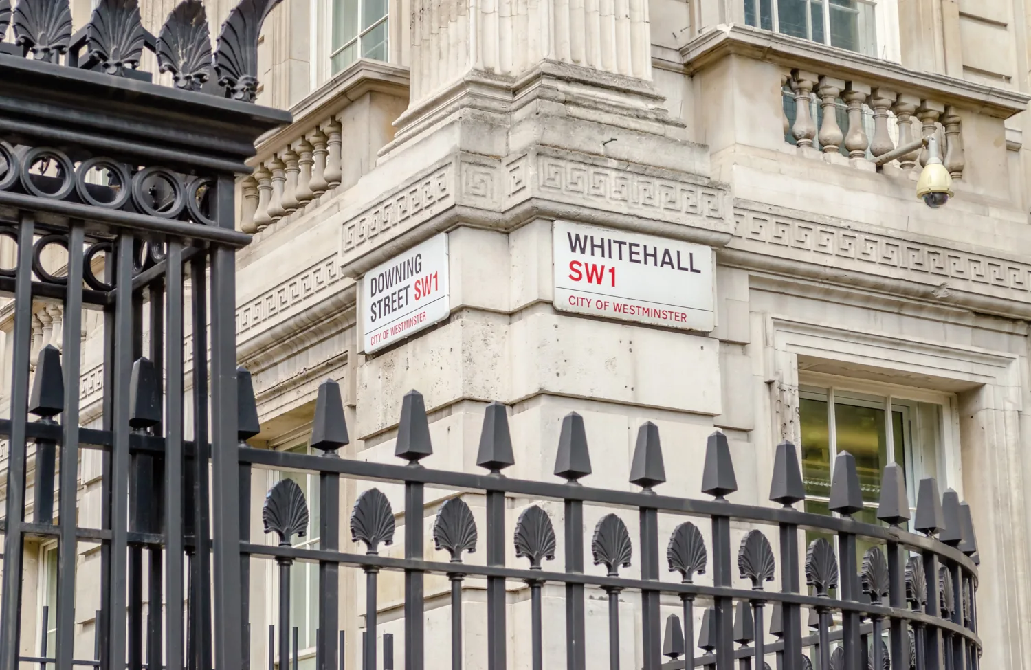 Street signs for Whitehall SW1 and Downing Street SW1 on a stone building corner in Westminster, with black iron railings in the foreground.