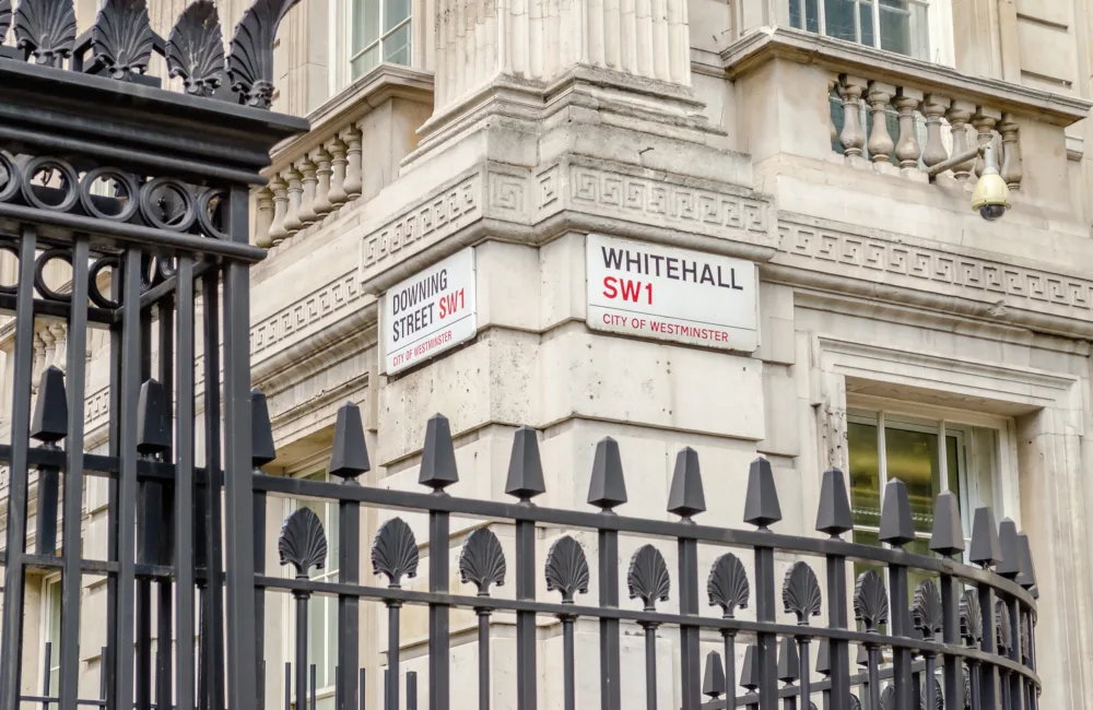 Street signs for Whitehall SW1 and Downing Street SW1 on a stone building corner in Westminster, with black iron railings in the foreground.