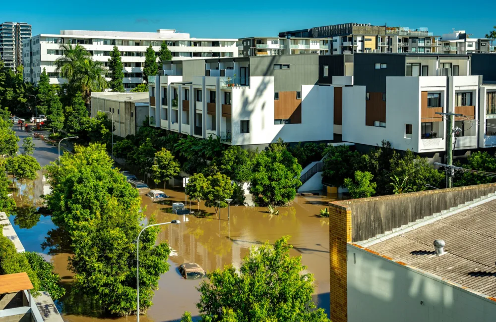 A flooded residential area with brown water submerging cars and partially covering trees. White and gray modern buildings are in the background, surrounded by greenery under a clear blue sky.