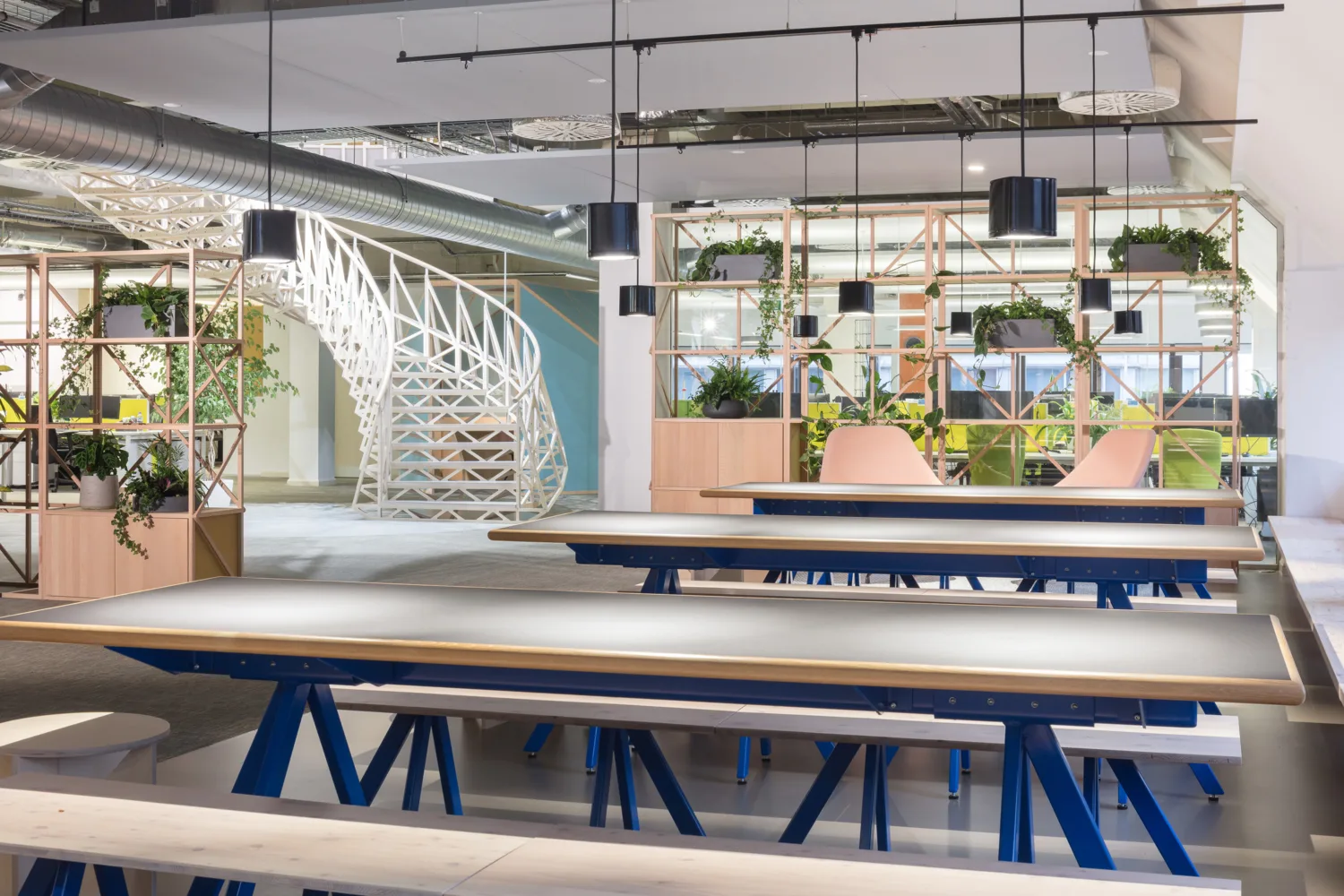 Workspace with long tables, bench seating, suspended lighting, exposed ductwork, planting and a white spiral staircase.