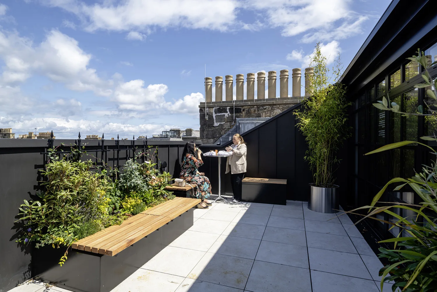 Rooftop terrace with planted beds, timber bench seating and two people at a table beneath a blue sky.