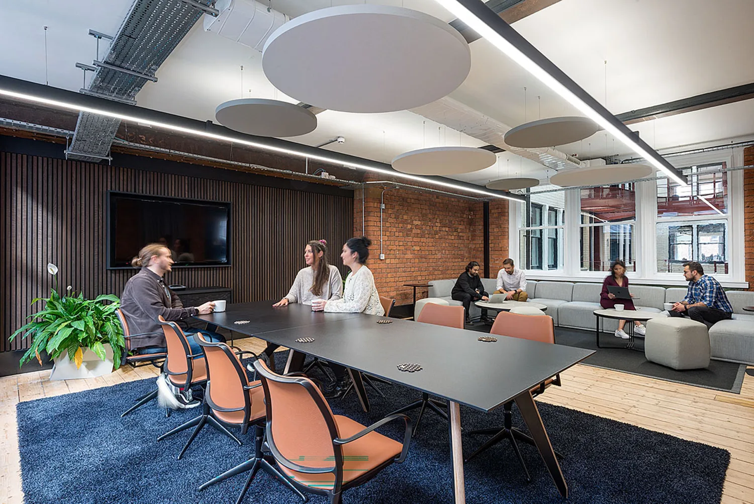 Workspace with a long table, lounge seating and several people, beneath suspended acoustic panels and linear lighting.