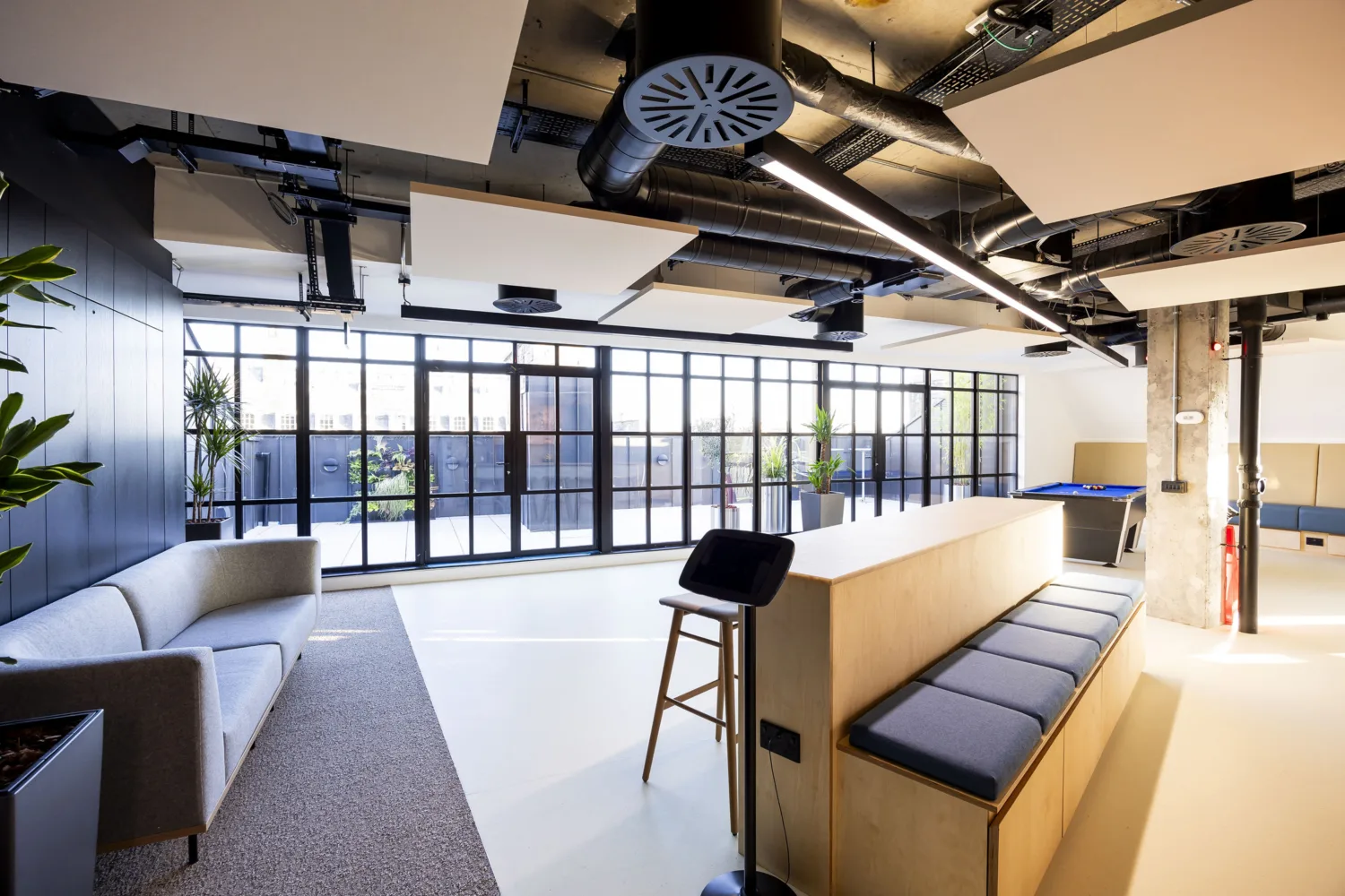Contemporary office interior with exposed ventilation ducts, acoustic ceiling panels, timber seating, sofas and floor-to-ceiling windows.
