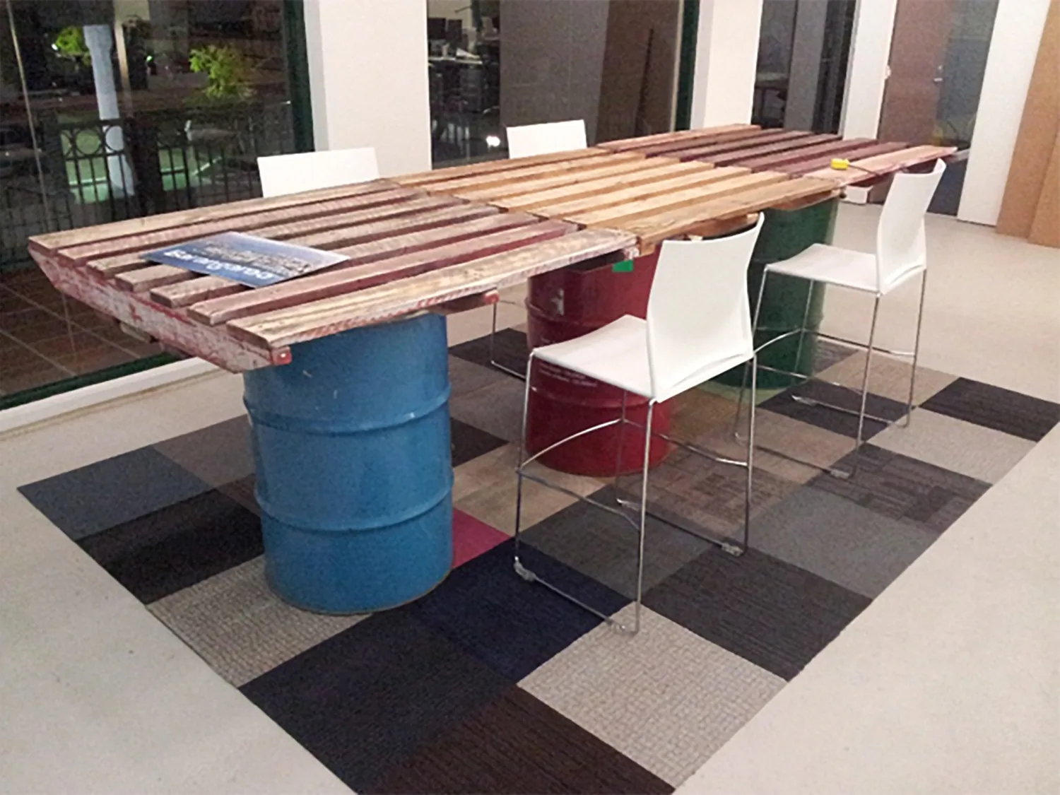 Long wooden slatted table supported by colourful metal barrels, surrounded by white chairs on a patchwork tiled floor.