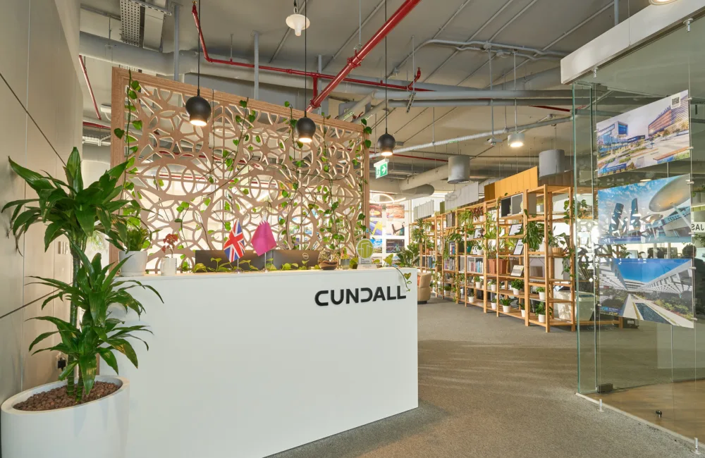 Modern office reception area with a white front desk labelled “CUNDALL”, indoor plants in pots, decorative wood screen with greenery, overhead exposed ceiling pipes, shelving units, glass partition with mounted project images, and carpeted flooring.