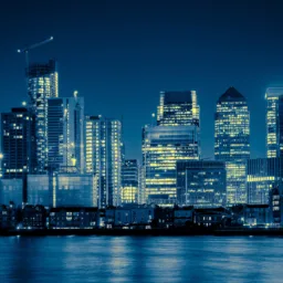 Night-time city skyline with illuminated high-rise office buildings and cranes, viewed across a calm river. Reflections of lights shimmer on the water, with dark blue sky and dense urban architecture in the background.