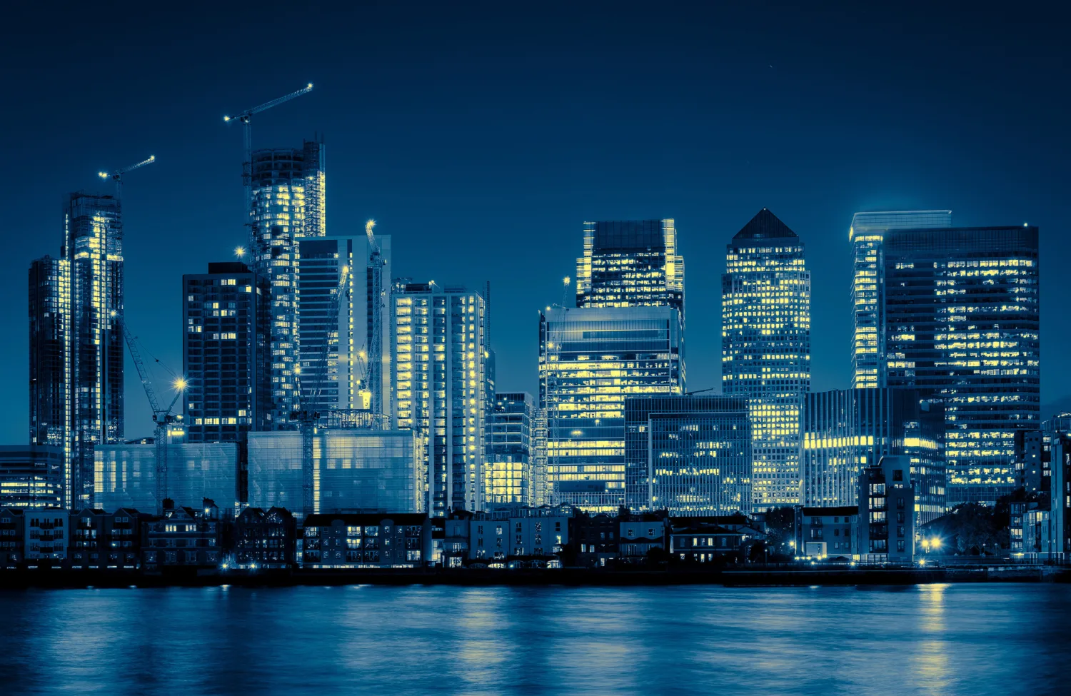 Night-time city skyline with illuminated high-rise office buildings and cranes, viewed across a calm river. Reflections of lights shimmer on the water, with dark blue sky and dense urban architecture in the background.