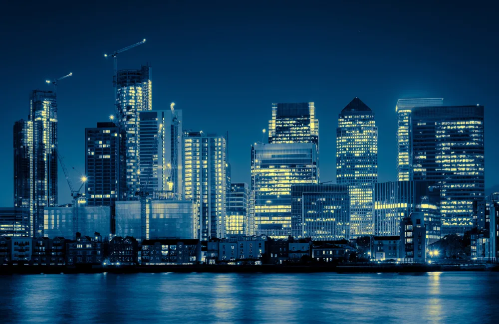 Night-time city skyline with illuminated high-rise office buildings and cranes, viewed across a calm river. Reflections of lights shimmer on the water, with dark blue sky and dense urban architecture in the background.