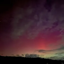 Red and green aurora illuminating clouds above a dark hillside under a star-filled night sky.