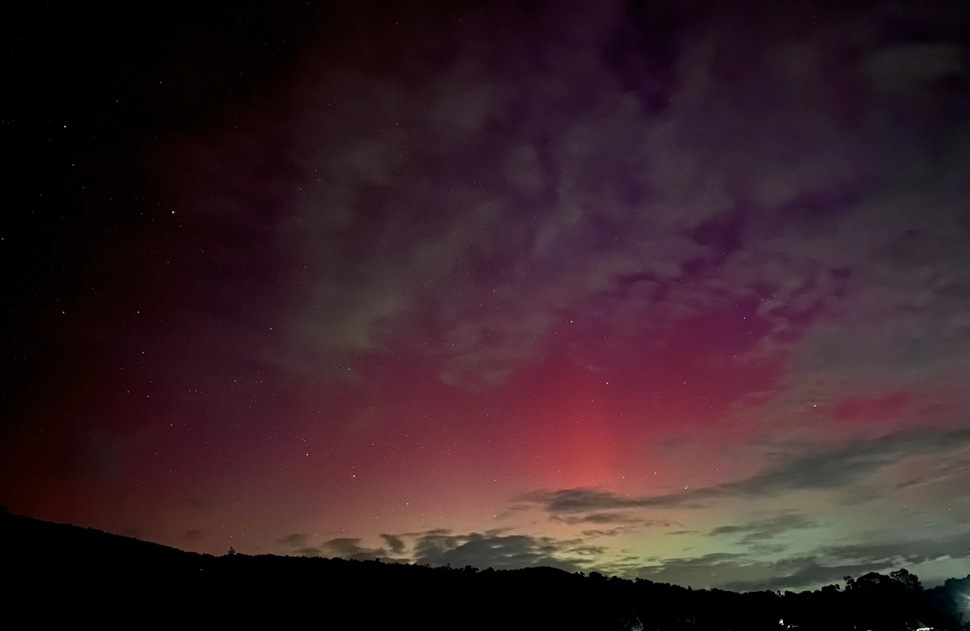 Red and green aurora illuminating clouds above a dark hillside under a star-filled night sky.