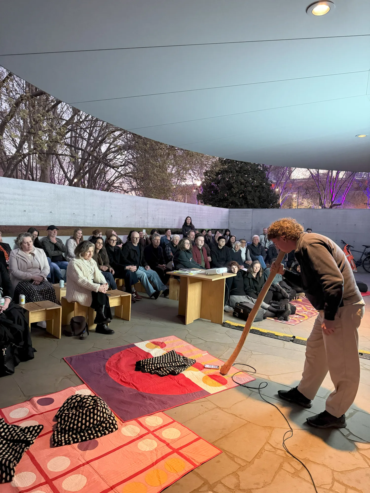 Audience seated around a performer playing a long wooden wind instrument on patterned floor mats beneath a curved canopy.