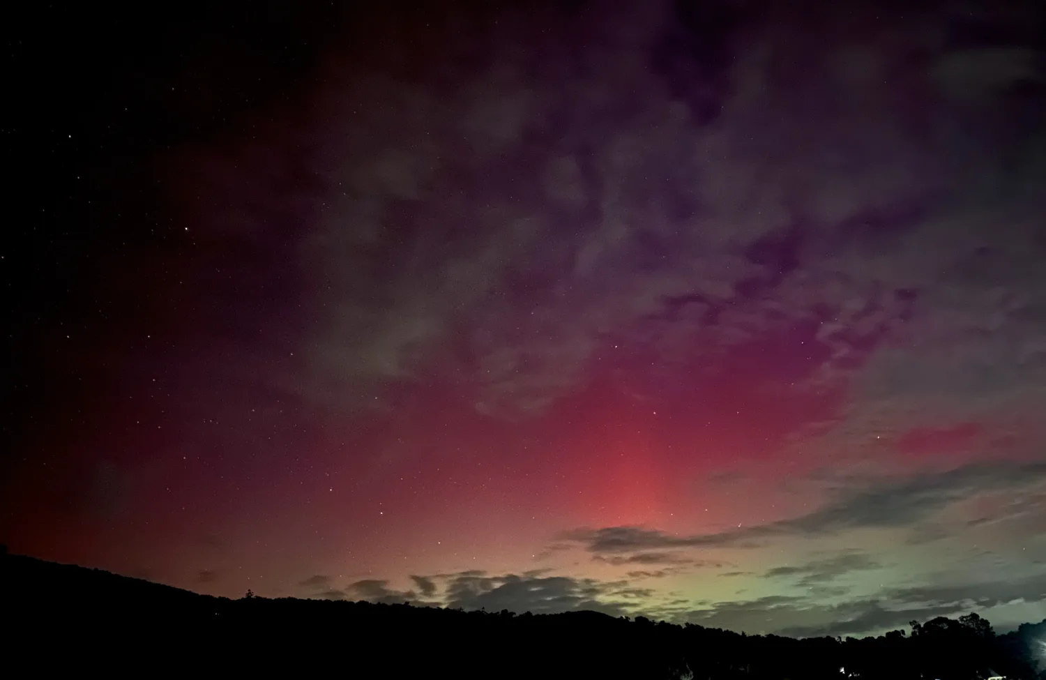 Red and green aurora illuminating clouds above a dark hillside under a star-filled night sky.