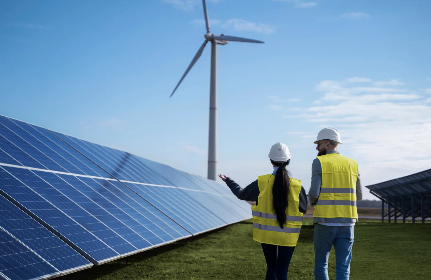 Two people wearing white hard hats and high-visibility yellow safety vests stand beside rows of blue solar panels on a grassy site. One person gestures toward the panels. A large wind turbine rises in the background beneath a partly cloudy sky.