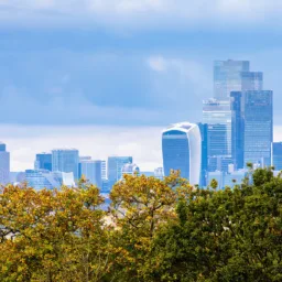 Panoramic view of the London skyline from a green park, with dense trees in the foreground and modern skyscrapers including The Shard, the Walkie Talkie, and The Gherkin in the distance beneath a cloudy blue-grey sky.