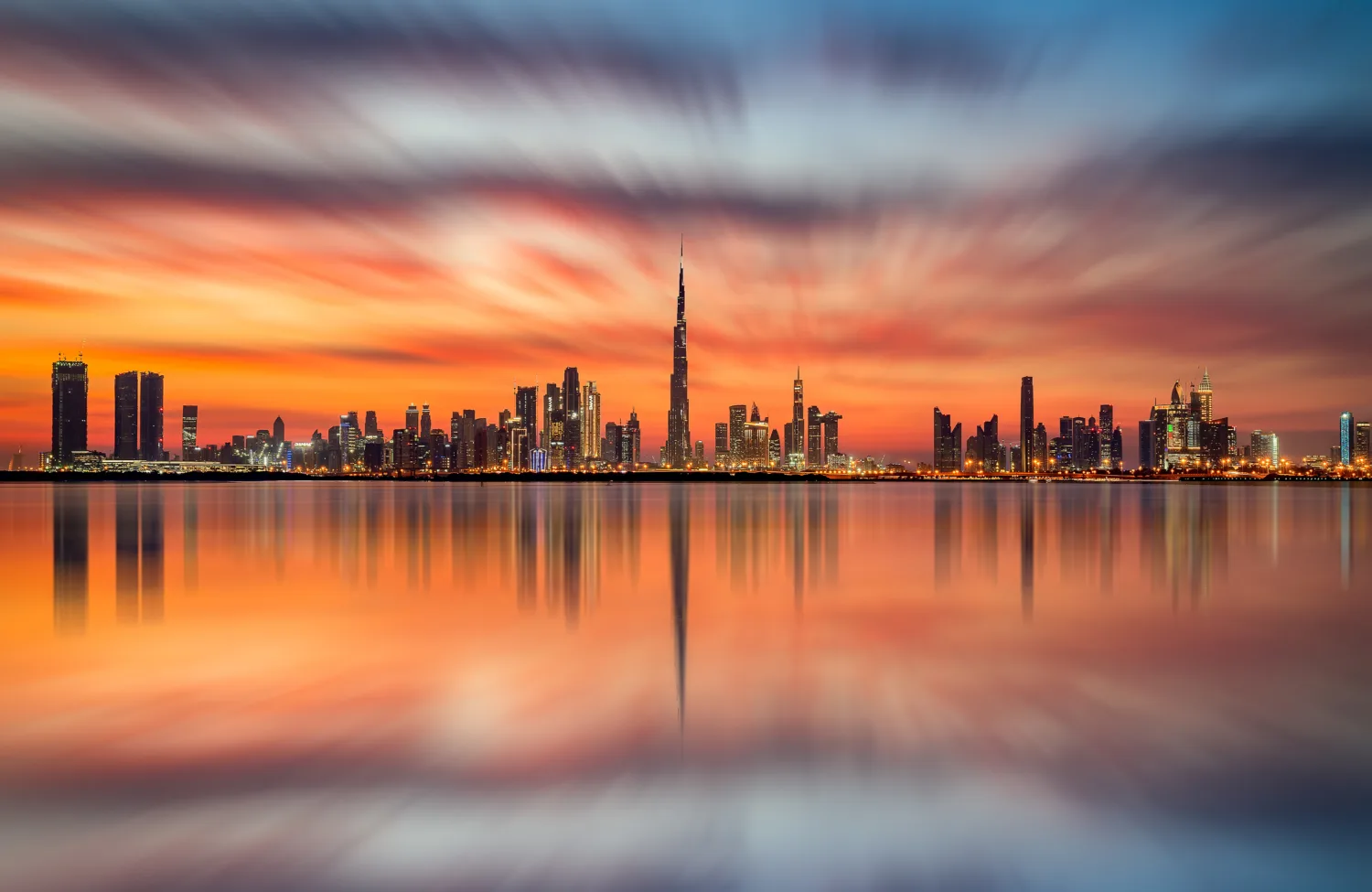 Dubai skyline at sunset with Burj Khalifa centred among high-rise buildings, reflected in calm water.