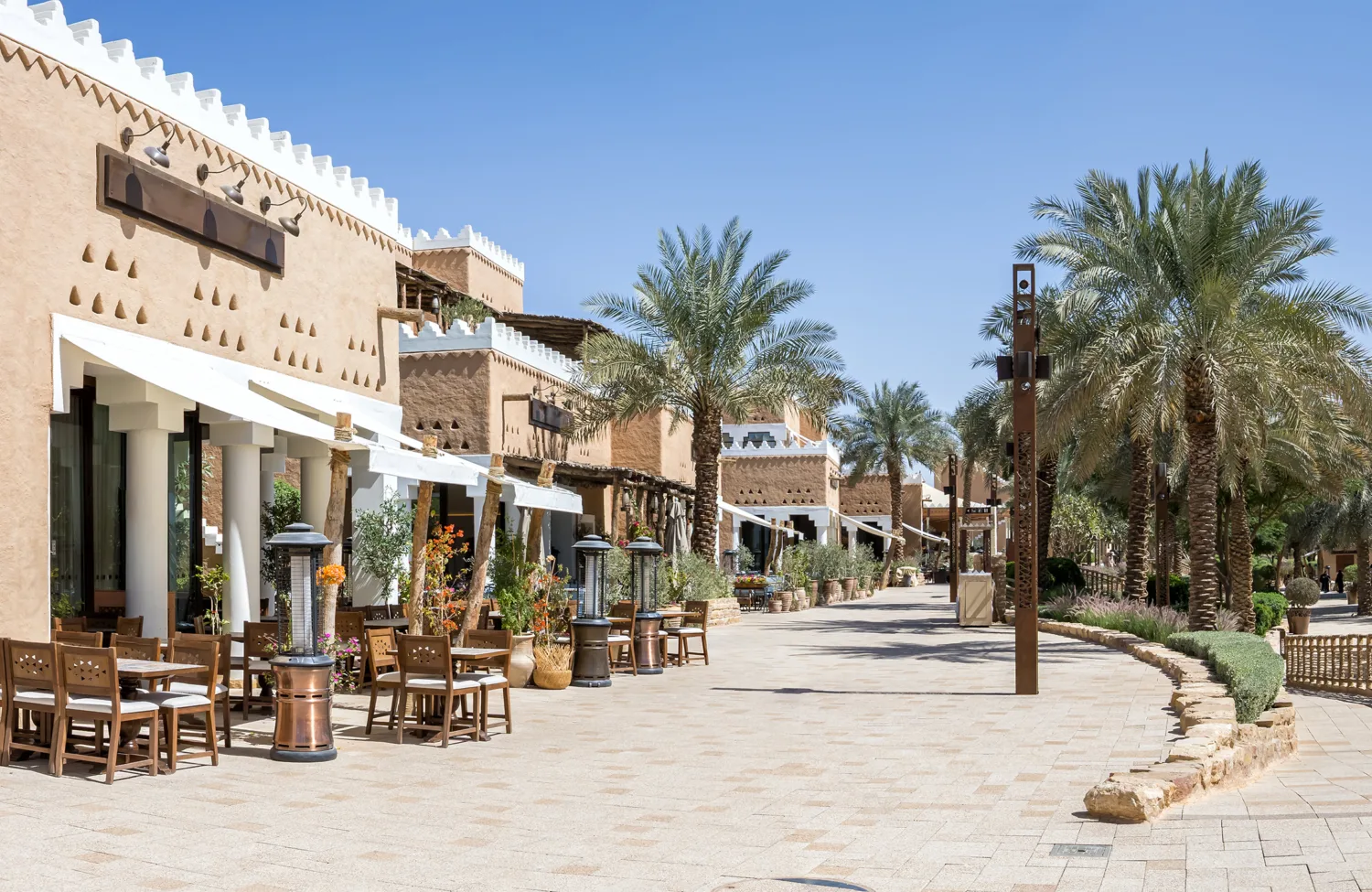 Pedestrian street lined with palm trees, outdoor seating and earth-toned buildings with shaded façades under a clear blue sky.