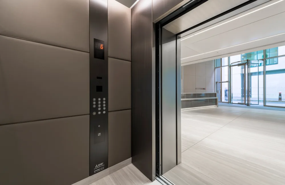 Interior of lift, controls panel, angling towards reception and entrance of an office building