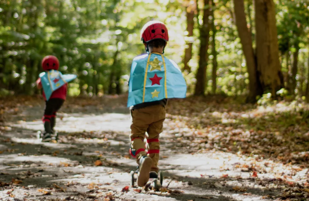 2 children on scooters wearing superhero capes on a woodland path