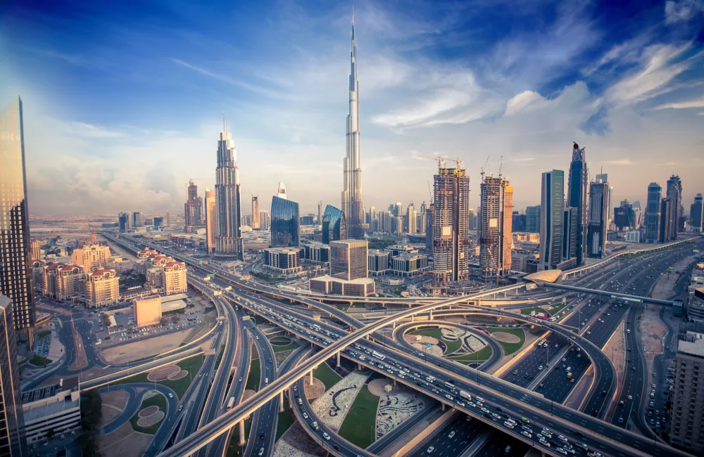 Dubai skyline at dusk with towers and construction shown behind a main road interchange